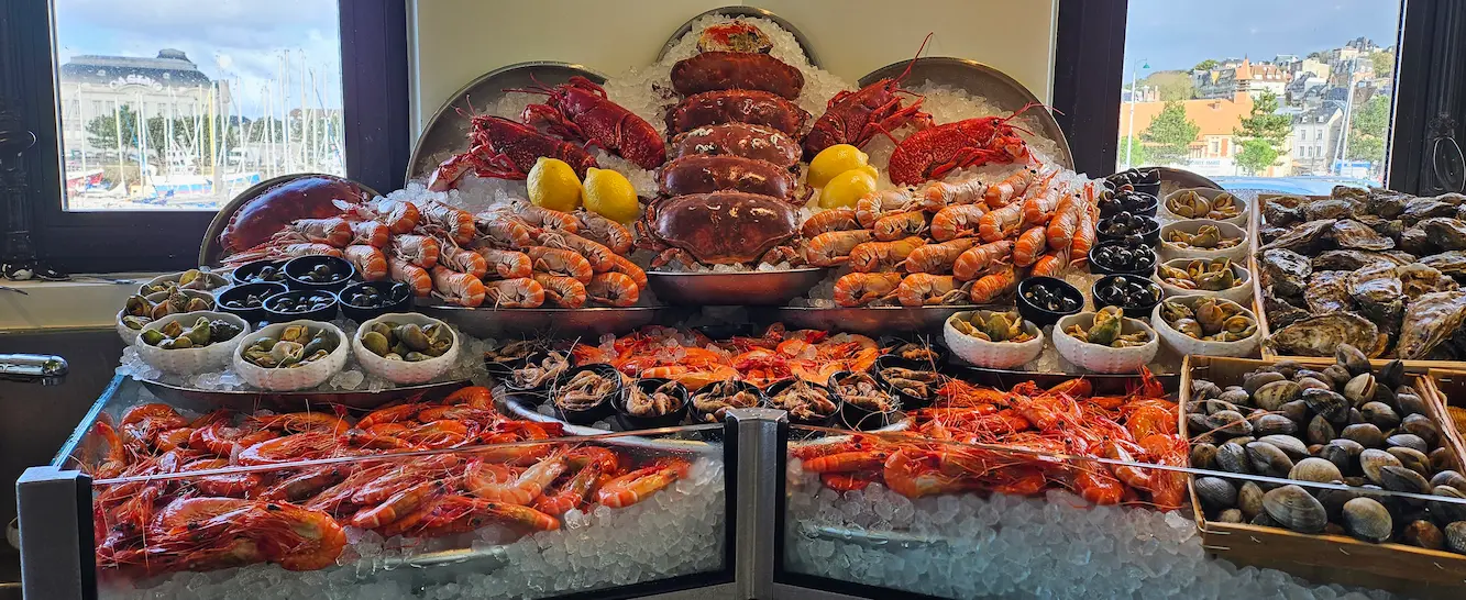 Étal de fruits de mer sur glace avec crevettes, crabes, homards, moules, palourdes et citrons devant des fenêtres donnant sur un port.