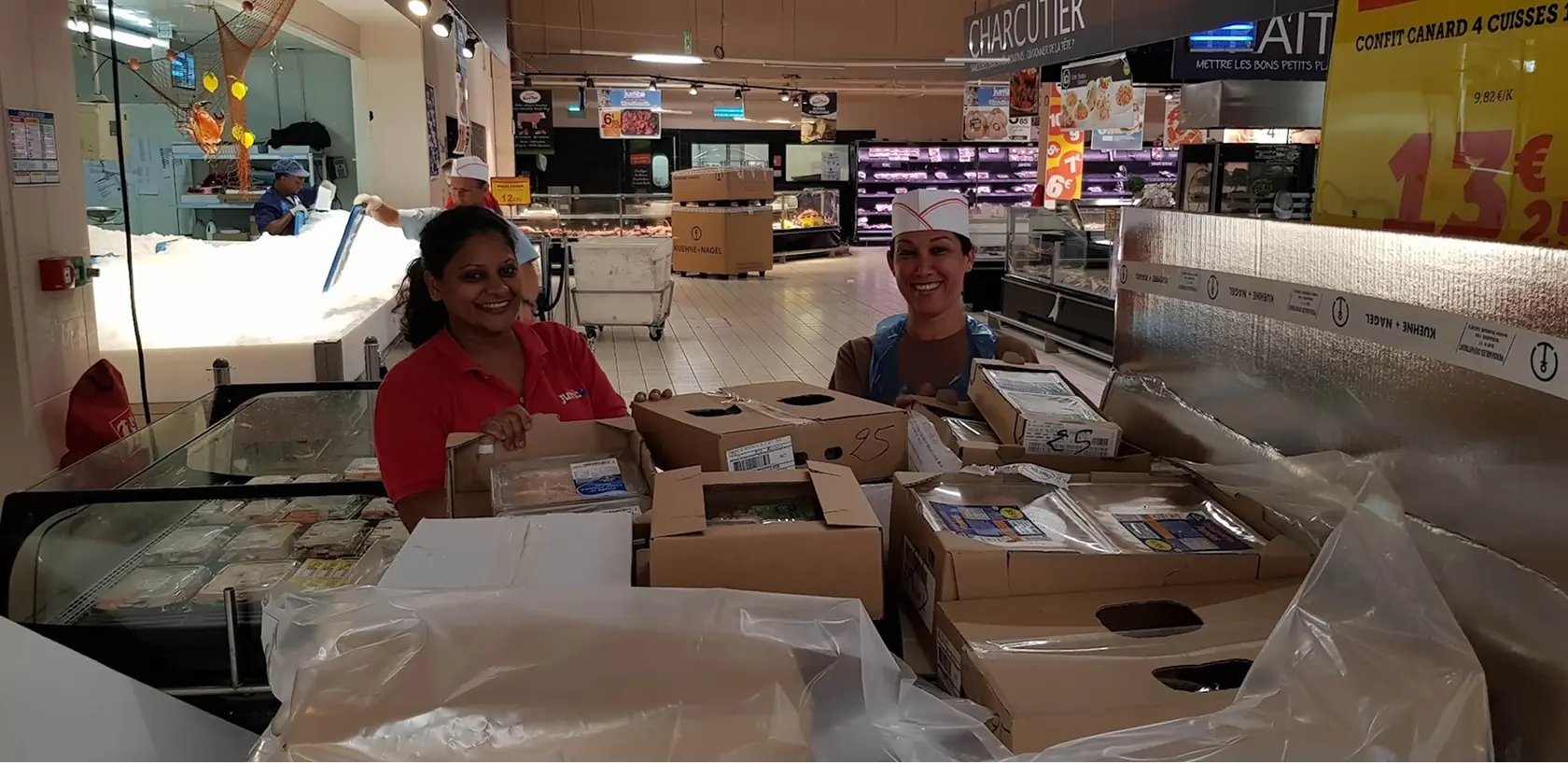 Deux employées souriantes derrière des cartons au rayon charcuterie d'un supermarché.