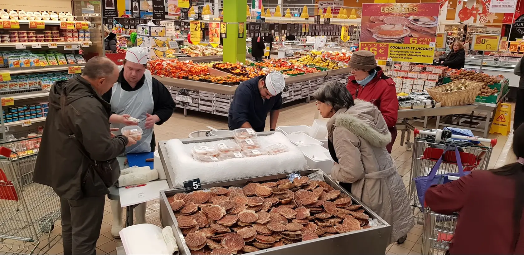 Un étal de coquilles Saint-Jacques vivantes dans un supermarché avec des clients et des employés en train de servir.