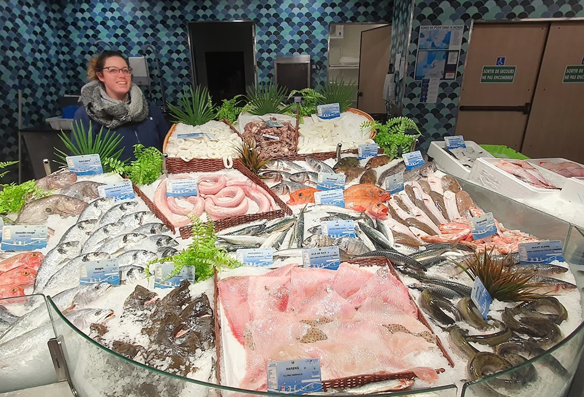 Femme souriante derrière un étal de poisson frais varié sur de la glace dans un marché couvert.