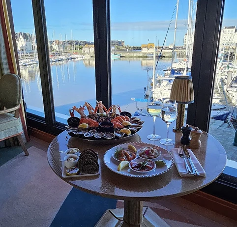 Table près d'une baie vitrée avec vue sur un port de plaisance, décorée avec un plateau de fruits de mer, deux verres de vin blanc, assiettes avec ceviche, et du pain.