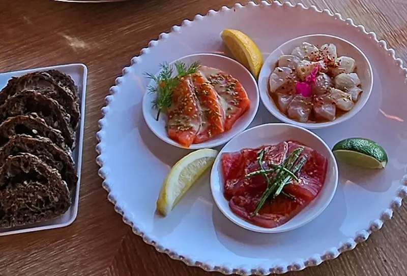 Trois petits bols contenant différents types de poissons crus marinés et des quartiers d'agrumes sont disposés sur une assiette blanche, à côté d'une assiette de pain noir tranché, sur une table en bois.