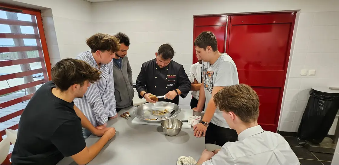 Un chef cuisinier montrant une technique de préparation culinaire à cinq jeunes attentifs autour d'une table grise en cuisine.