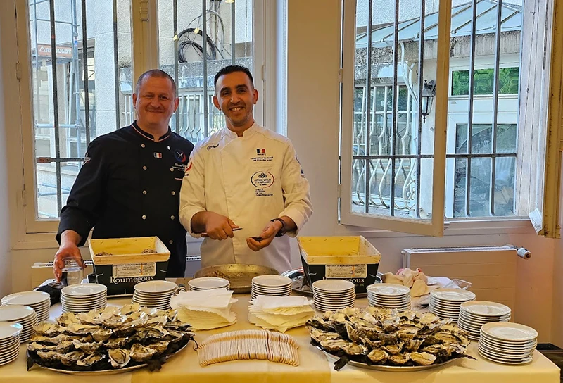 Dave Vitré et un chef en tenue professionnelle, debout derrière une table avec des plateaux d’huîtres ouvertes, des assiettes empilées et des serviettes dans une pièce éclairée par une fenêtre ouverte.