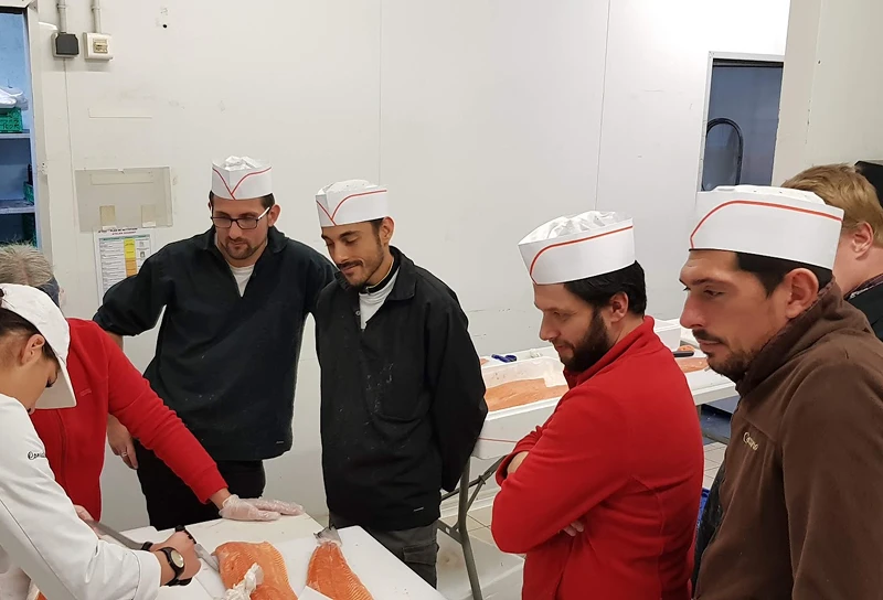 Un groupe d'hommes portant des casquettes blanches observe une personne en uniforme montrant comment découper un filet de saumon sur une table blanche.