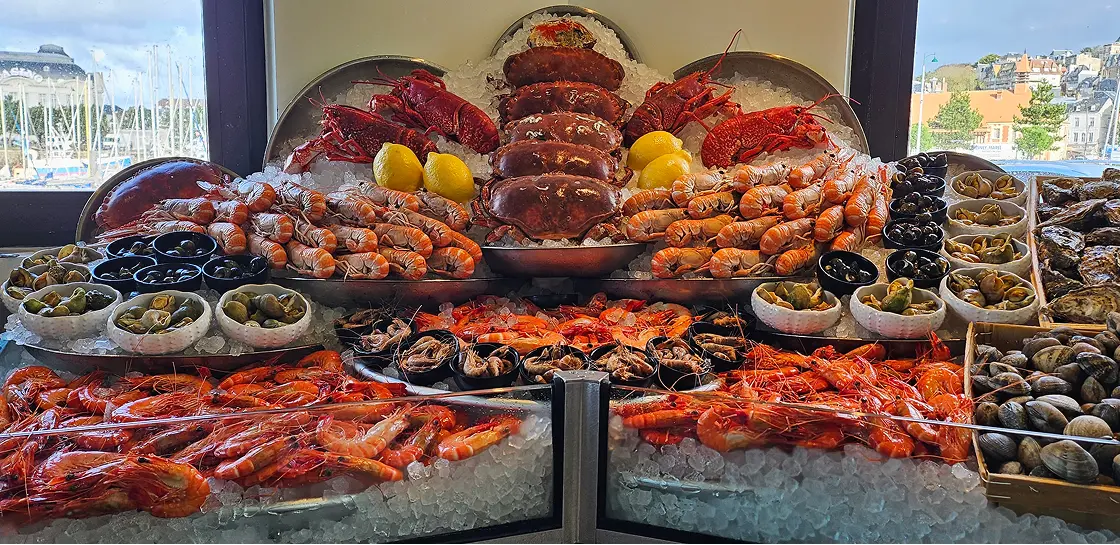 Grande étalage de fruits de mer frais sur glace comprenant homards, crabes, crevettes, escargots de mer, palourdes et huîtres devant une fenêtre avec vue sur un port.