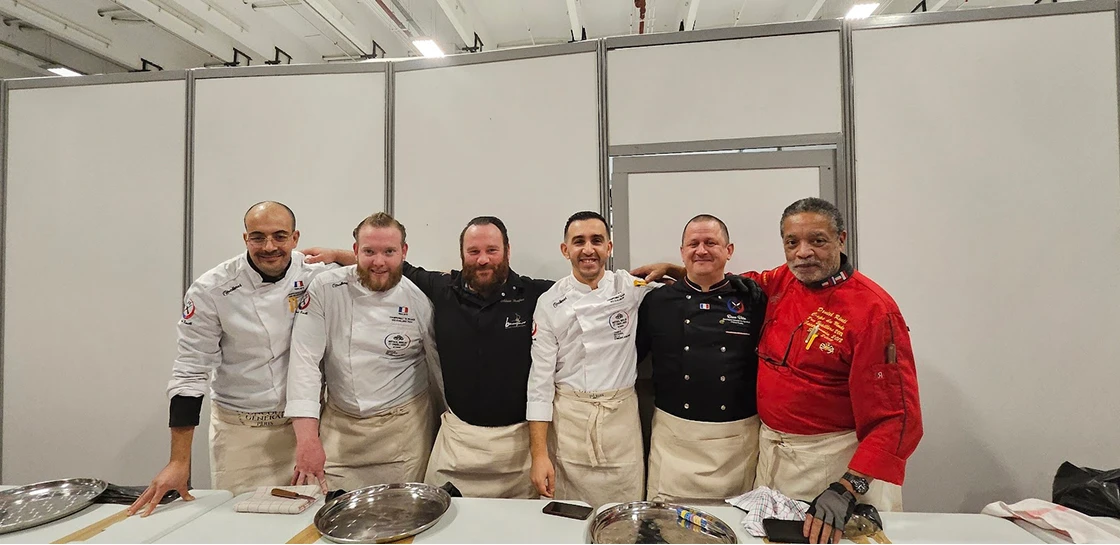 Six chefs souriants portant des uniformes avec tabliers posent ensemble derrière une table avec plateaux en métal dans un cadre intérieur.
