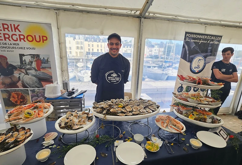 Homme souriant debout derrière une table de fruits de mer frais, avec des plateaux d'huîtres, crevettes et coquillages dans une tente au bord d'un port.