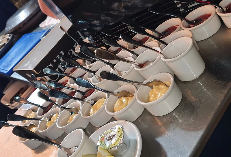 Rangée de petits ramequins blancs contenant différentes sauces comme de la moutarde et des confitures, chacun avec une petite cuillère, sur une table en métal.