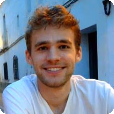 Smiling young man with short curly brown hair wearing a white T-shirt outdoors near a white building.