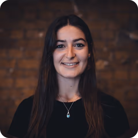 Smiling woman with long dark hair wearing a black top and a teardrop-shaped pendant necklace against a blurred brick wall background.