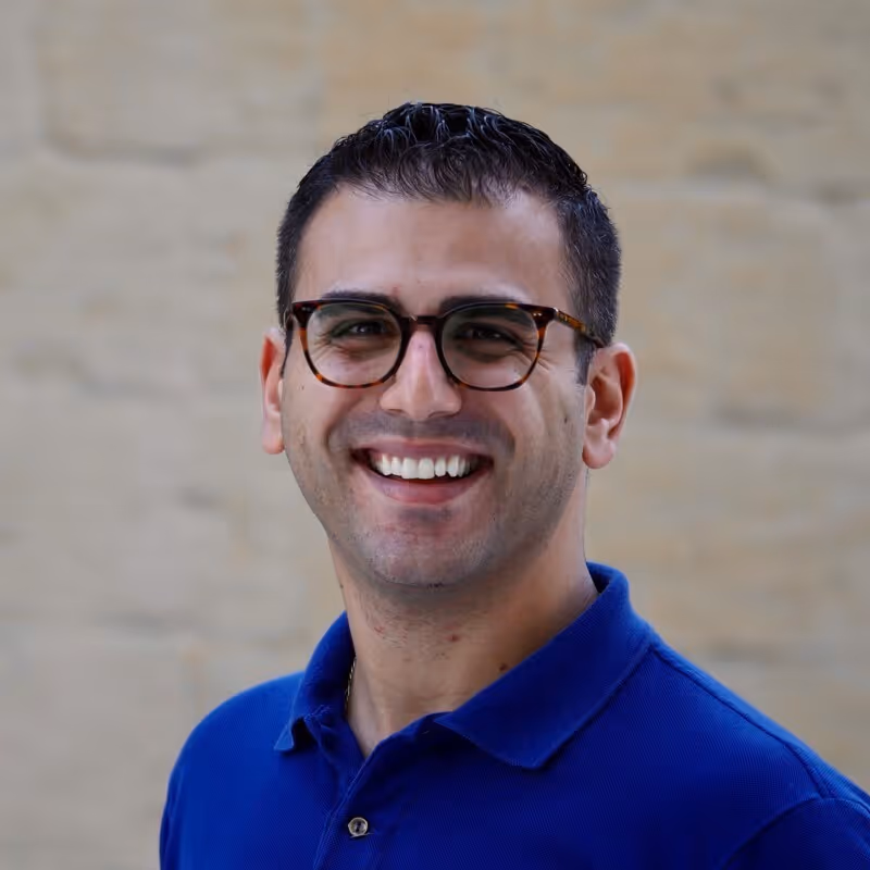 Smiling man with short dark hair and glasses wearing a blue polo shirt in front of a beige brick wall.