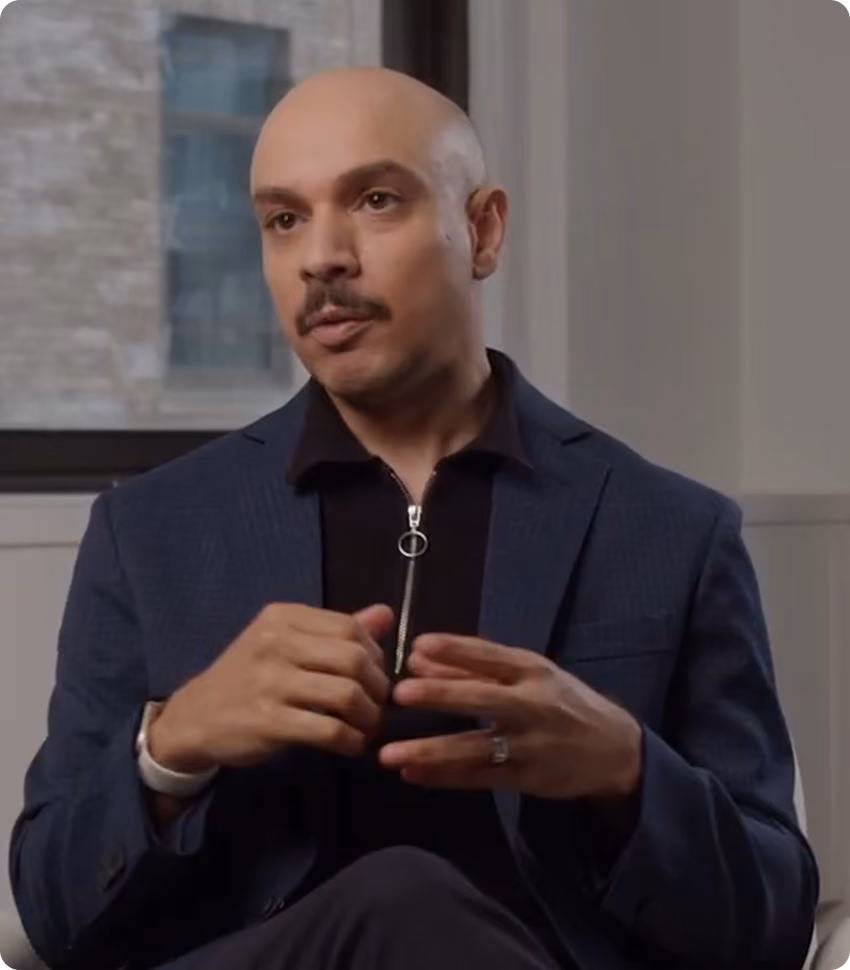 Bald man with mustache wearing a dark blazer and black shirt, gesturing with his hands while seated.