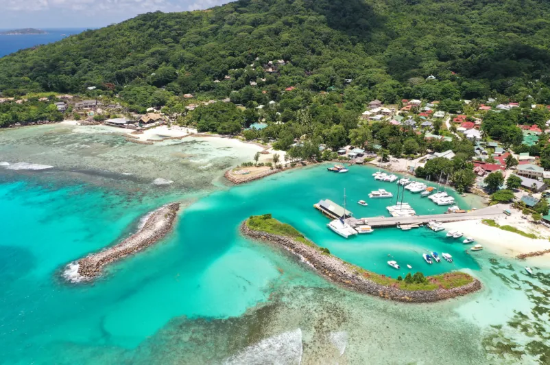 Aerial view of La Digue ferry terminal in Seychelles, showing turquoise waters, protective rock barriers, boats at anchor, and the lush island landscape—key connection point for Cat Rose and SeyFerry travelers.
