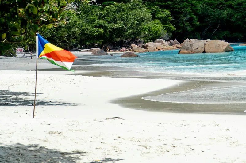 Seychelles national flag on a white sand beach with turquoise water, granite boulders, and lush green vegetation in the background.