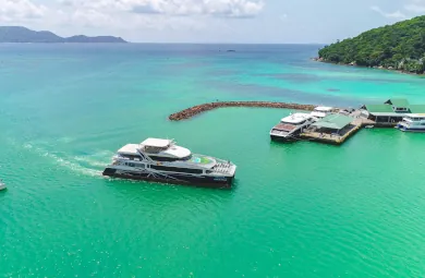 Cat Cocos high-speed ferry approaching the jetty at Baie Sainte Anne, Praslin, with turquoise waters and lush green hills in the background.