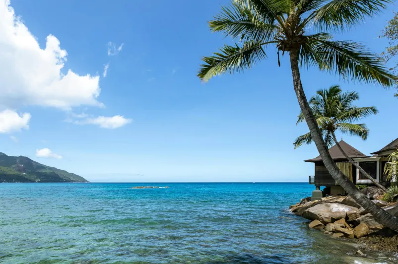 View of the Seychelles coastline with clear blue waters, a tropical beach villa, and palm trees overlooking the ocean—typical scenery along the Mahe to Praslin ferry route.