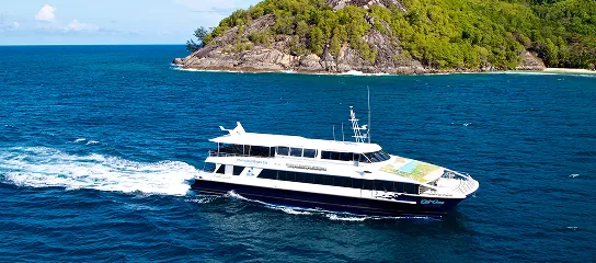 Cat Cocos ferry cruising past a rocky, forested coastline in Seychelles under a clear blue sky.
