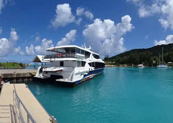 Cat Cocos ferry docked at the jetty in Seychelles, with turquoise water, a mountainous island in the background, and a shuttle van nearby.