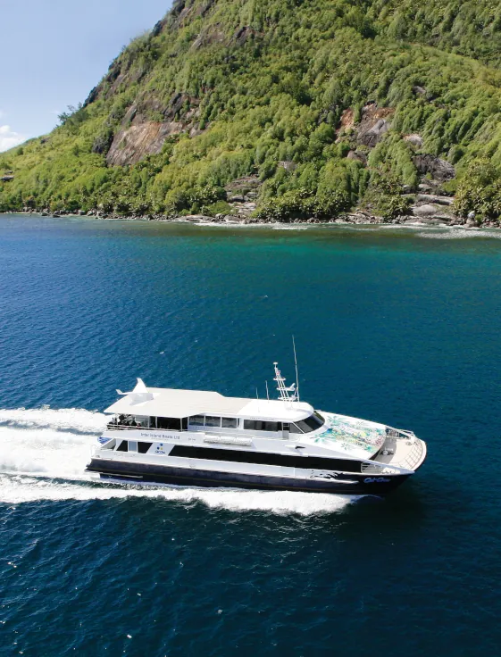 White ferry boat cruising on deep blue water alongside a lush green hillside coast in Seychelles.