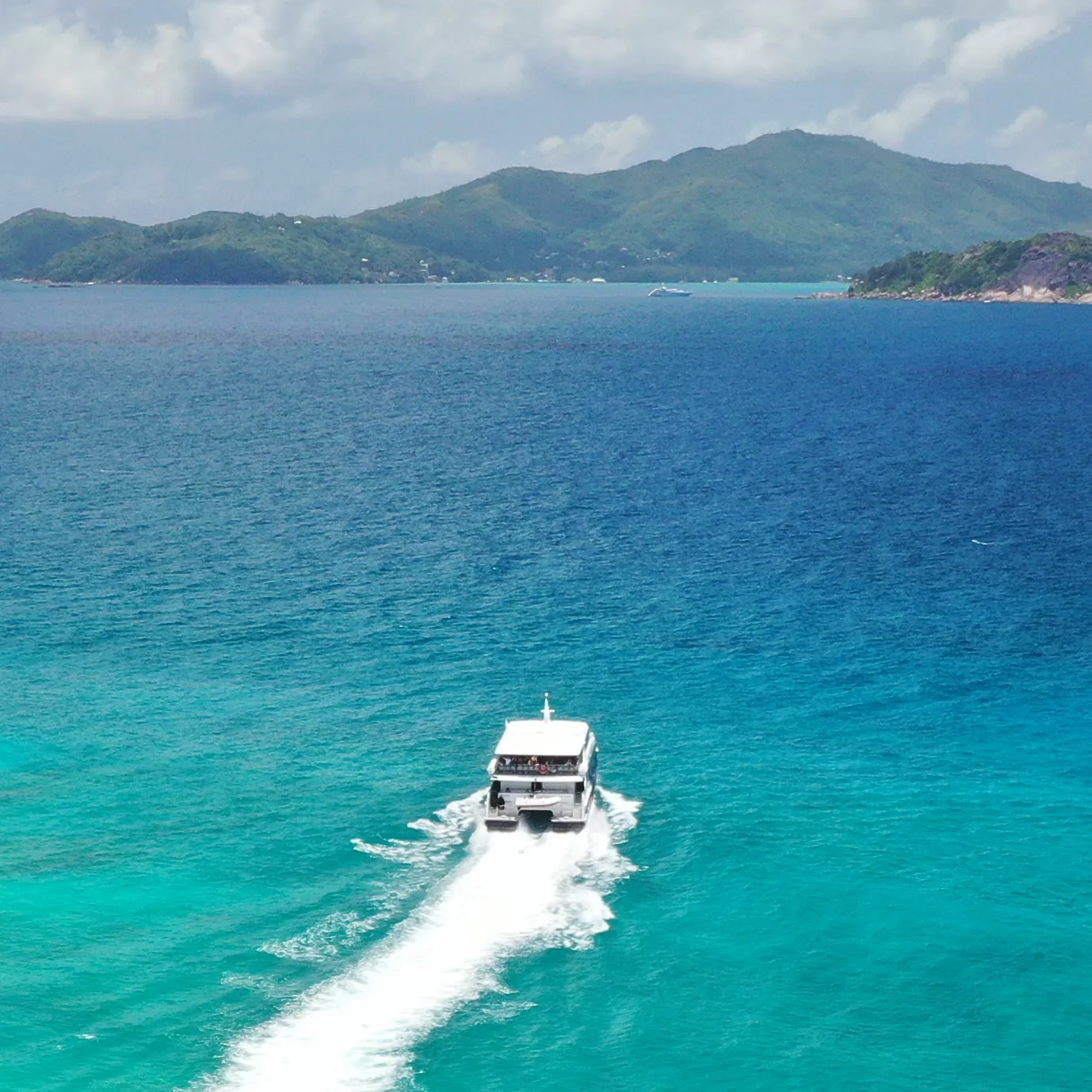 White boat speeding on clear turquoise water with green mountains under a cloudy sky in Seychelles.
