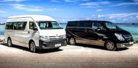 A white van and a black van parked side by side near a seaside under a partly cloudy sky in Seychelles.