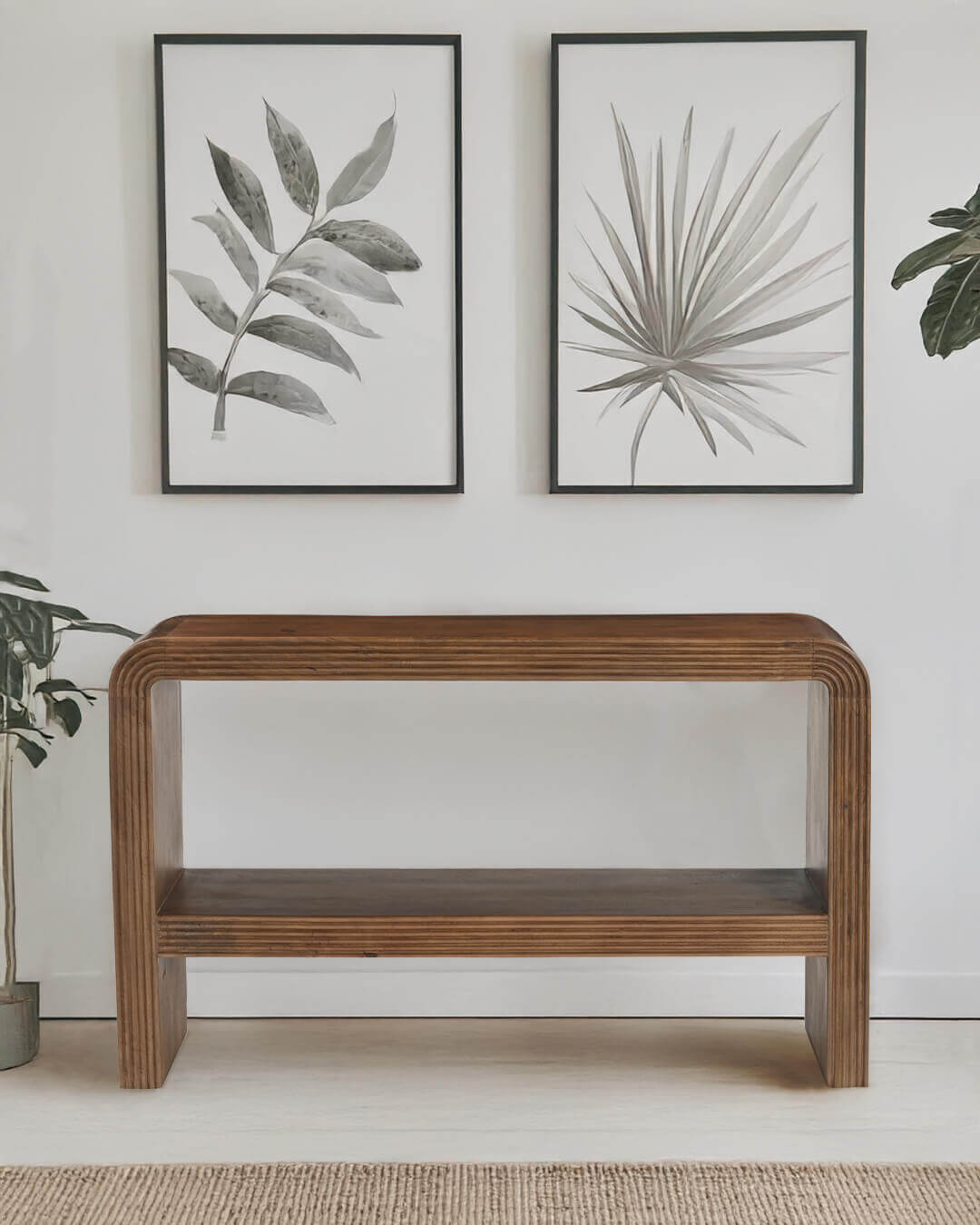 Minimalist wooden console table with rounded edges and a lower shelf, placed against a white wall with two monochrome botanical prints above it and green plants on either side.