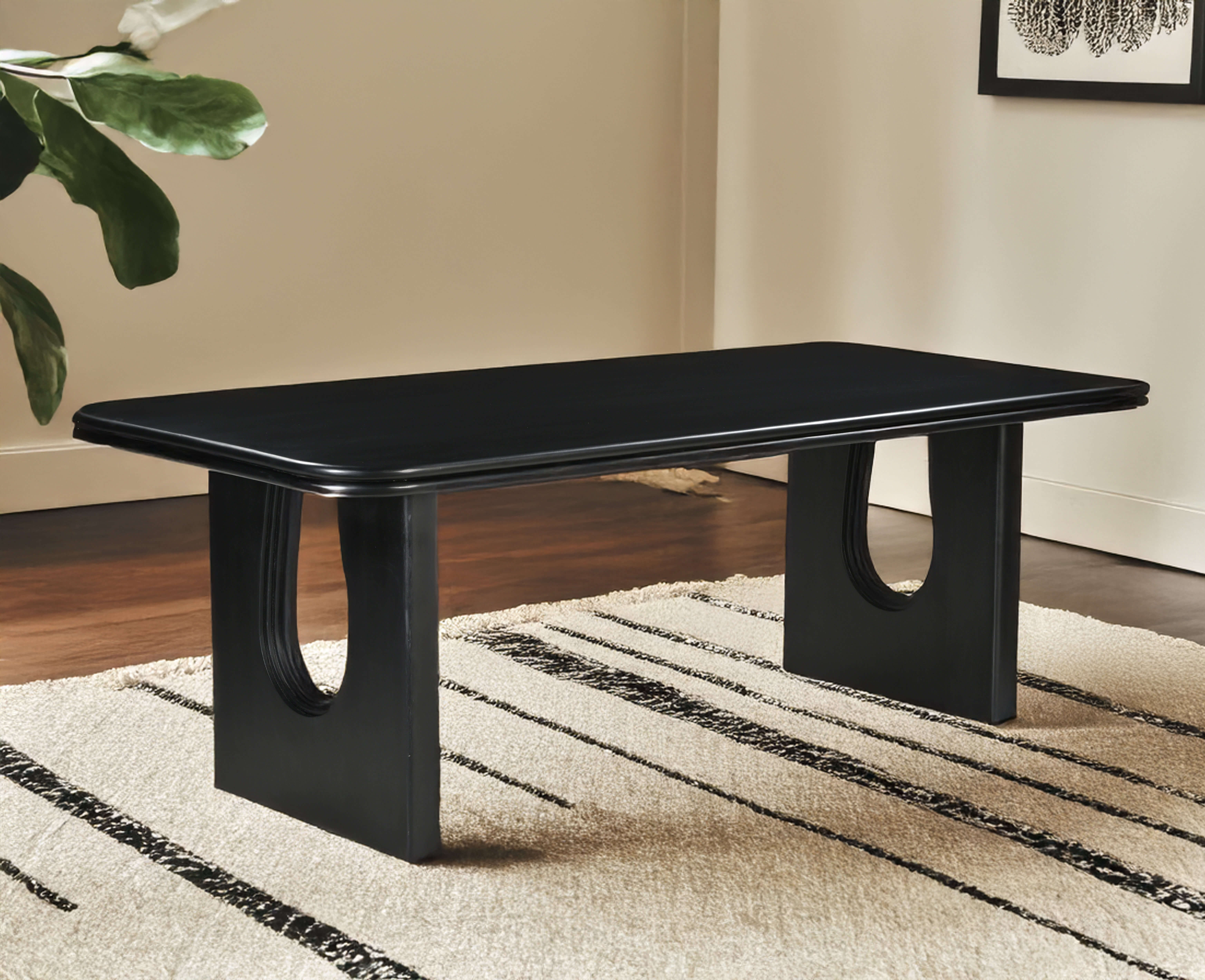 Modern black rectangular dining table with curved cut-out legs, placed on a cream and black striped rug in a minimal living space with wooden floors and neutral walls.
