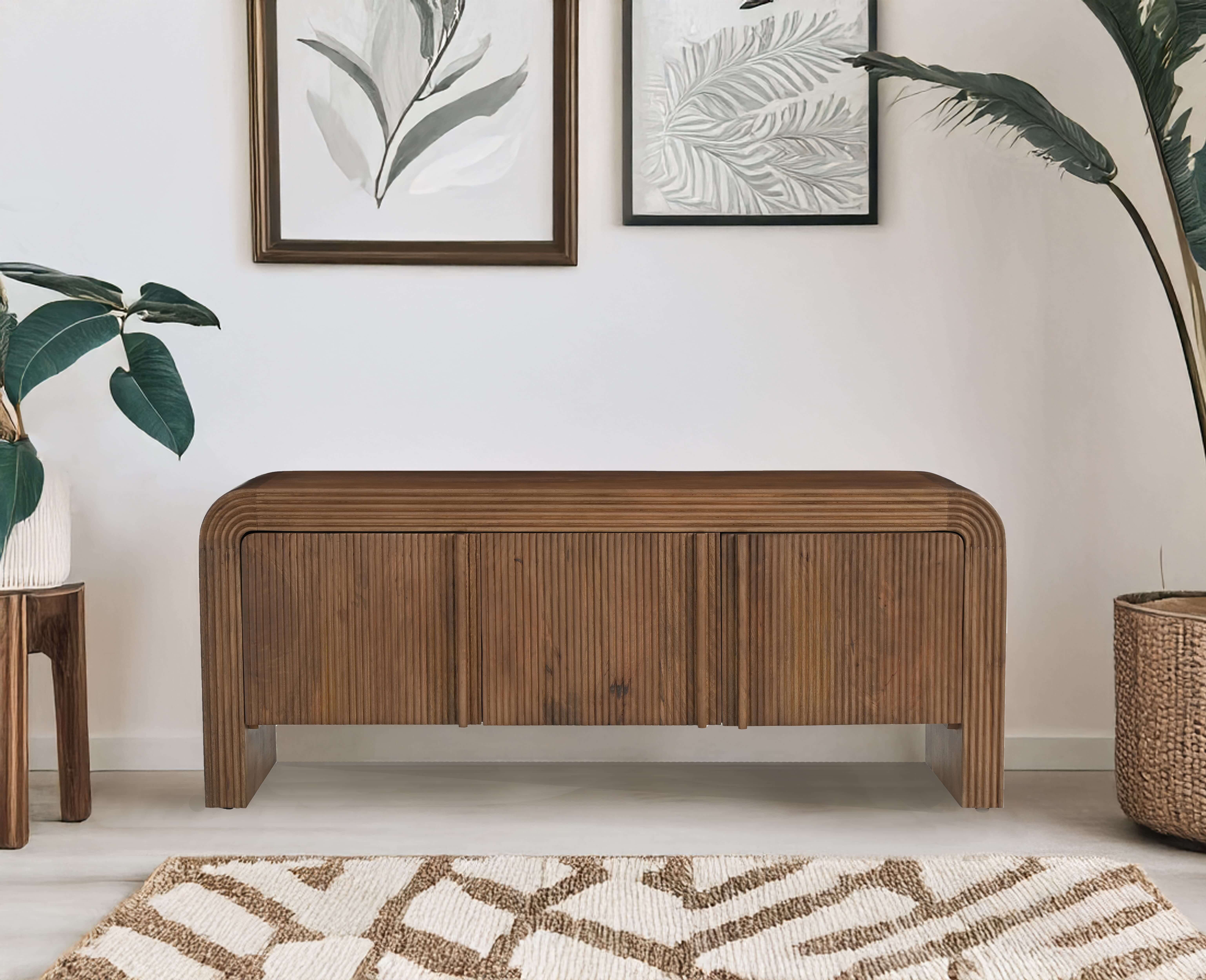 Tall wooden bookshelf with open shelves and a closed cabinet at the bottom, featuring a carved door design, placed against a neutral wall with a snake plant and jute rug for decor