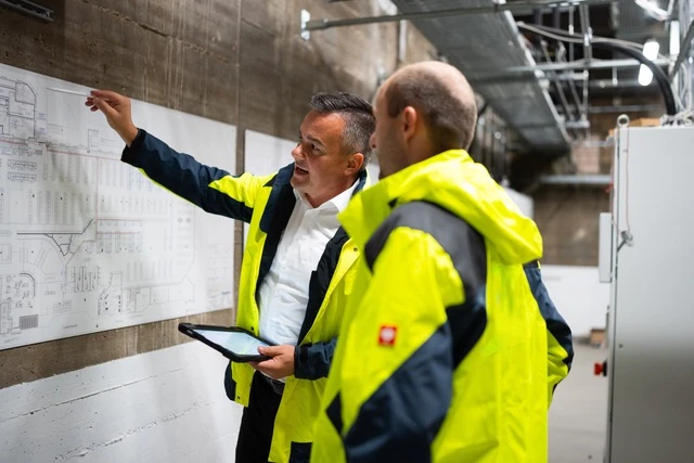 Zwei Männer in gelb-schwarzen Arbeitsjacken betrachten und zeigen auf technische Pläne an einer Wand in einem Industriegebäude