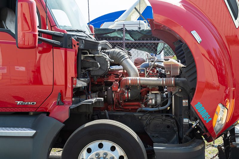 Red Kenworth T680 semi-truck with hood open, showing detailed engine components during repair, with technician inspecting under bright daylight.