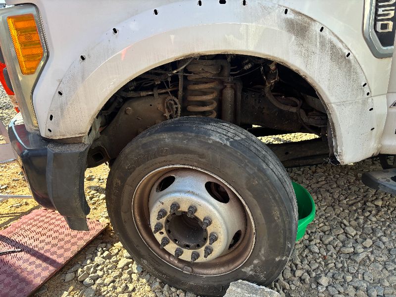 A white heavy-duty truck with the front fender removed, exposing the suspension coil spring and wheel assembly during repair work on a gravel surface.