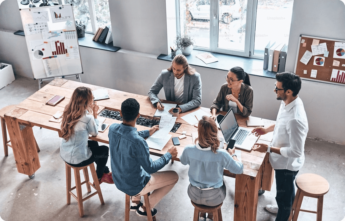 A group of business professionals sitting around a wooden table in a modern office, collaborating on documents and laptops during a brainstorming meeting.