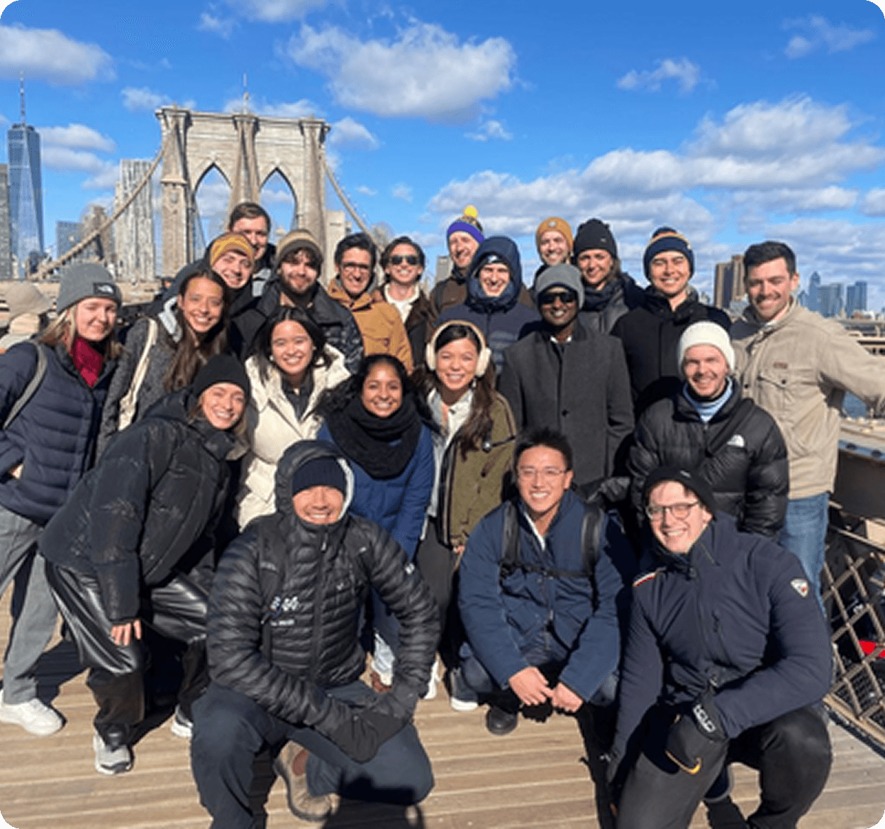 A diverse group of young professionals posing for a team photo on the Brooklyn Bridge in New York City on a sunny winter day, wearing warm jackets and smiling against a skyline background.
