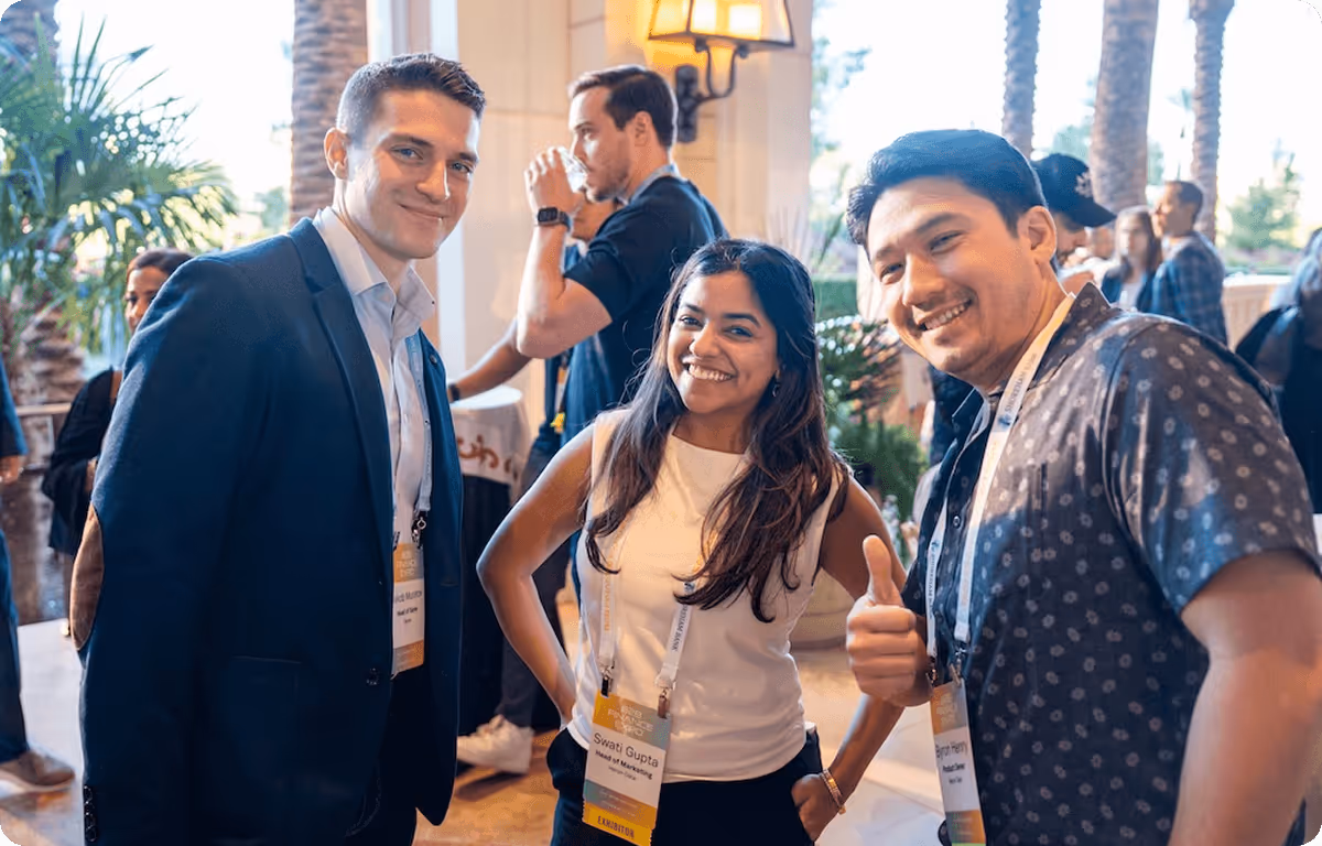 Three professionals smiling and posing at a networking event, wearing conference badges, with other attendees and palm trees in the background.