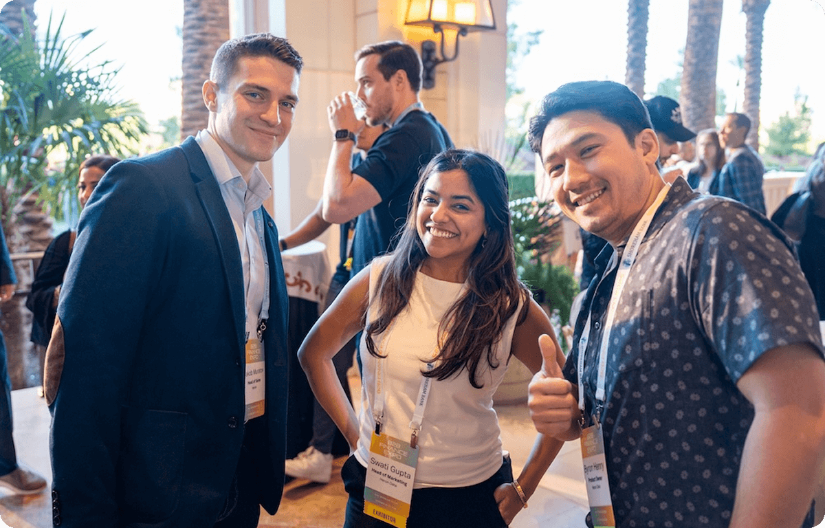 Three professionals smiling and posing at a networking event, wearing conference badges, with other attendees and palm trees in the background.
