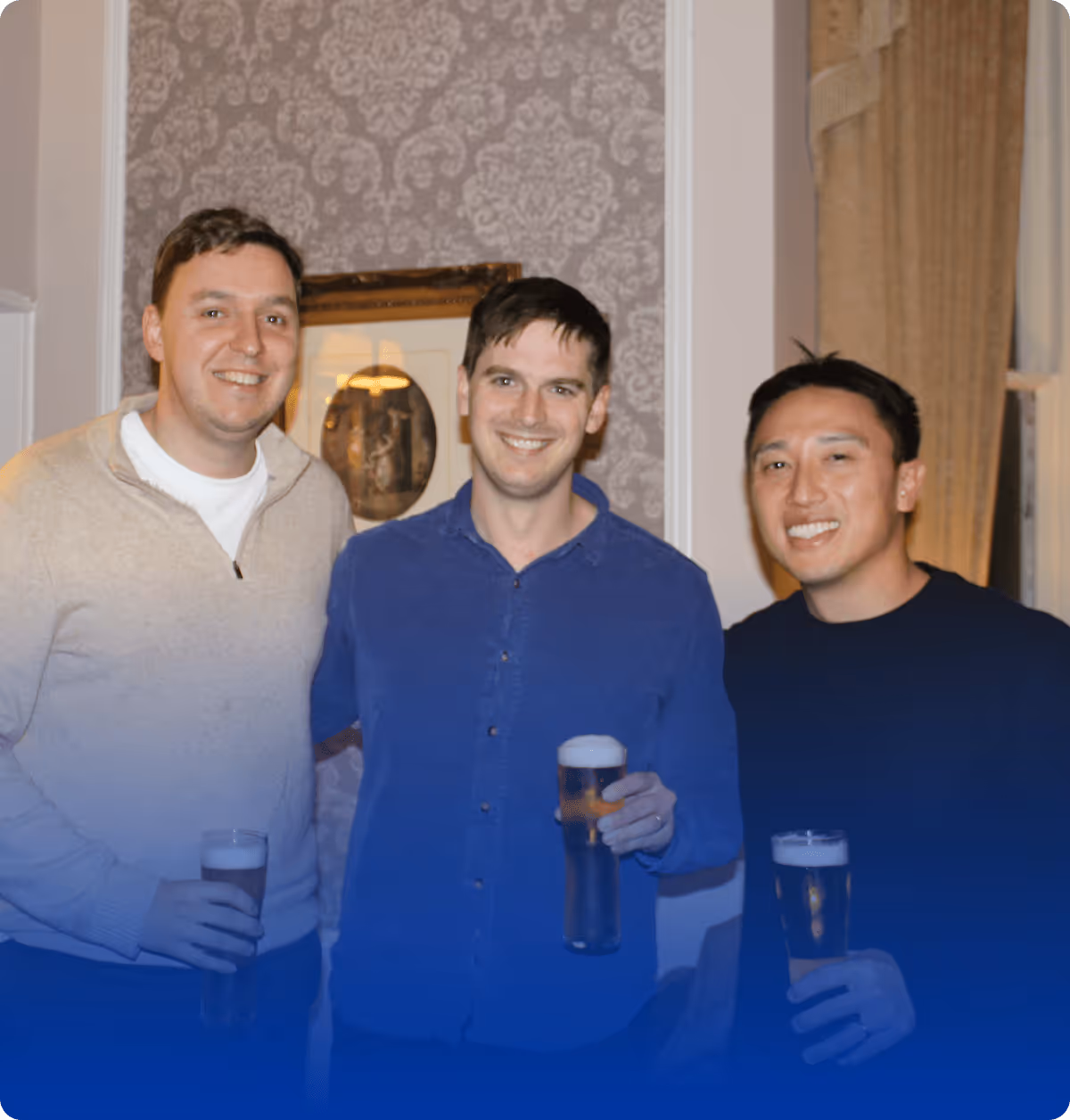 Three smiling men standing indoors in a warmly lit room, each holding a pint of beer, posing together in front of patterned wallpaper and framed artwork