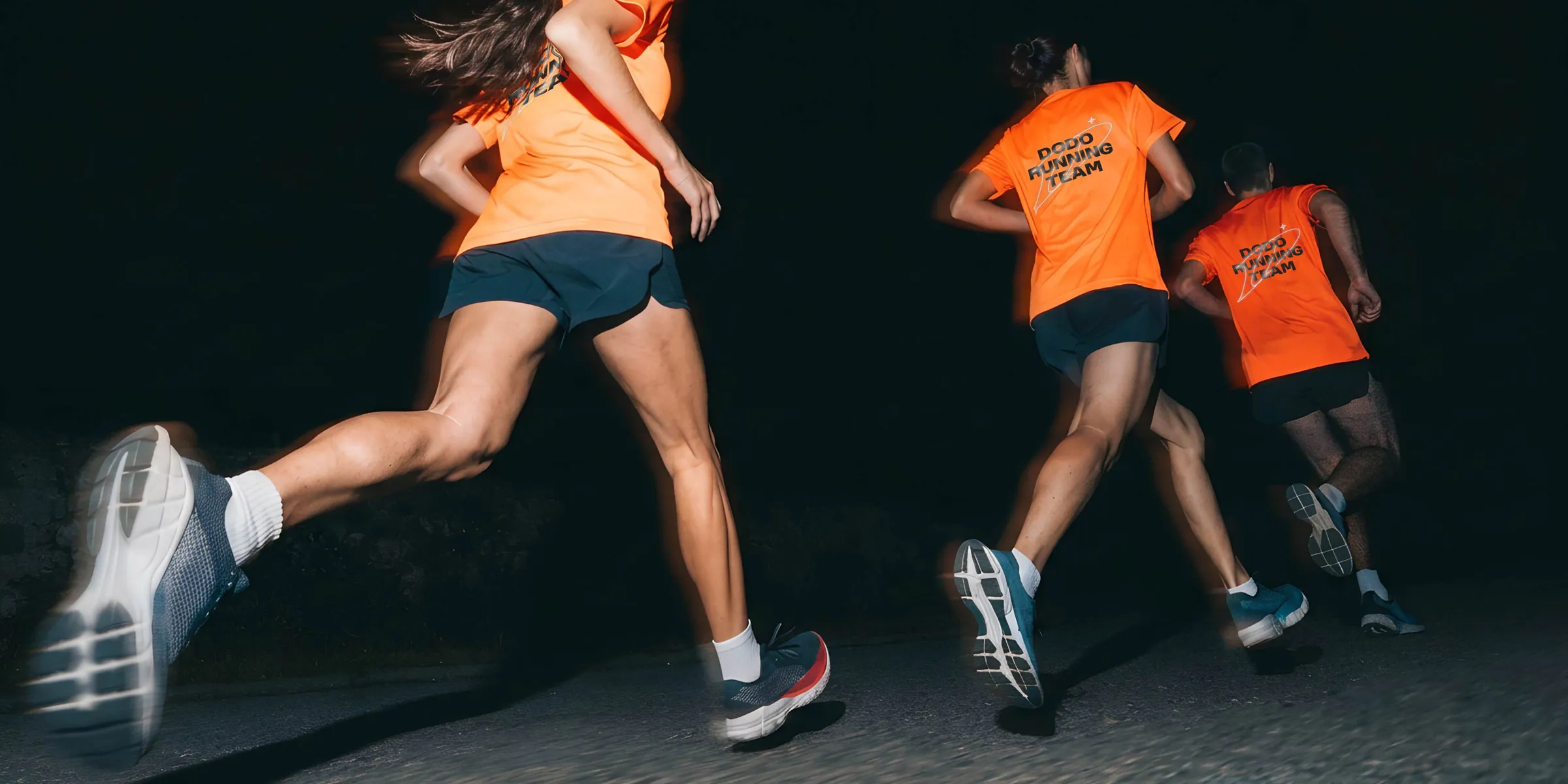 Три бегуна в оранжевых футболках с надписью DODO Running Team бегут ночью по дороге.