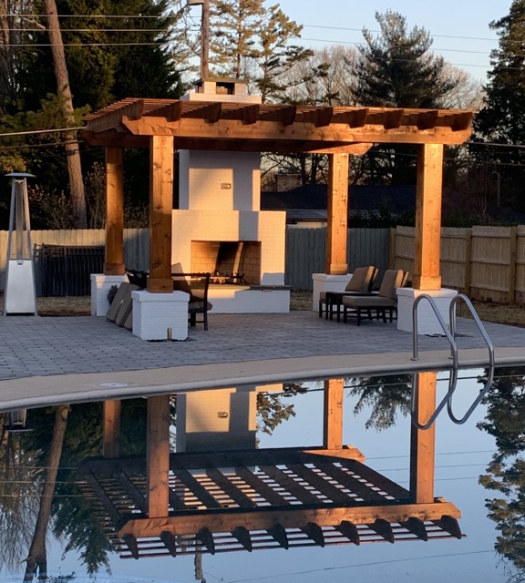 Outdoor patio with a wooden pergola and white brick fireplace reflected in a still swimming pool.