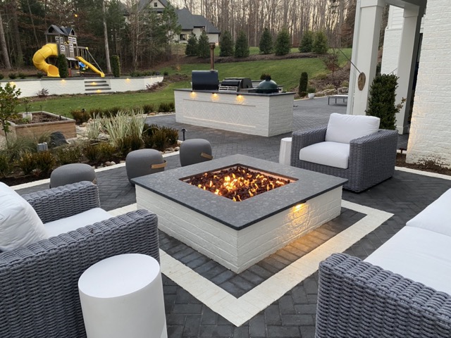 Modern outdoor seating area with a square gas fire pit surrounded by gray wicker chairs and white cushions on a paved patio.