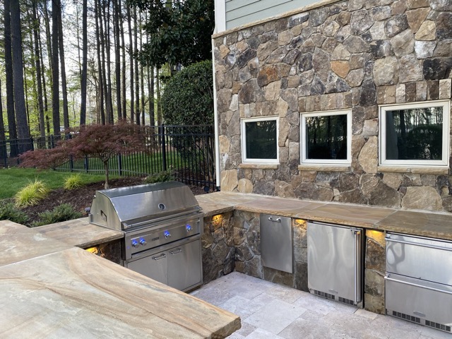 Outdoor kitchen with stone walls, stainless steel grill, fridge, and cabinets under a wooden countertop surrounded by trees.