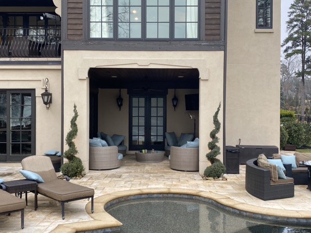 Outdoor patio with cushioned lounge chairs, circular wicker sofas with blue pillows under a covered area, and a curved swimming pool in the foreground.