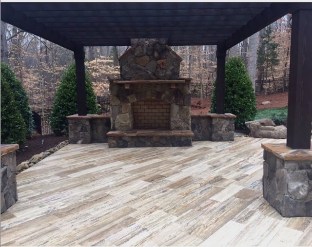 Outdoor stone fireplace with seating under a dark wooden pergola on a light wood-textured stone patio surrounded by bushes and trees.