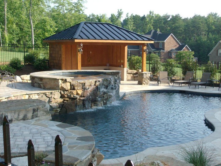 Outdoor swimming pool with an attached elevated hot tub featuring a stone waterfall edge, surrounded by lounge chairs and a wooden gazebo with a black metal roof.