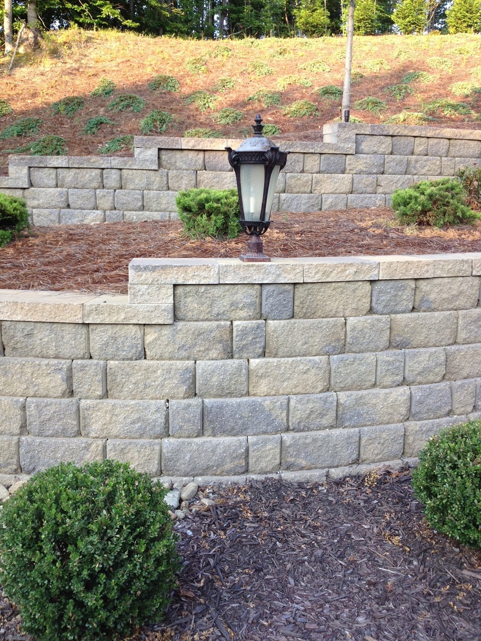 Stone retaining wall with tiered landscaping, small shrubs, pine straw mulch, and a black decorative outdoor lamp.