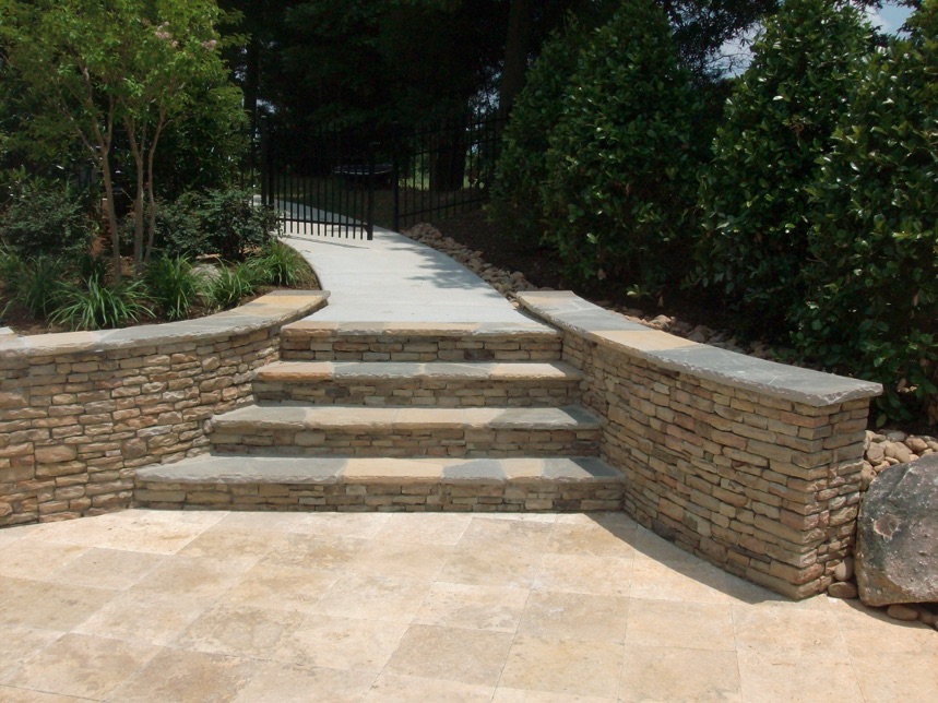 Stone steps leading to a white concrete pathway with curved stacked stone walls on both sides, surrounded by greenery and bushes.