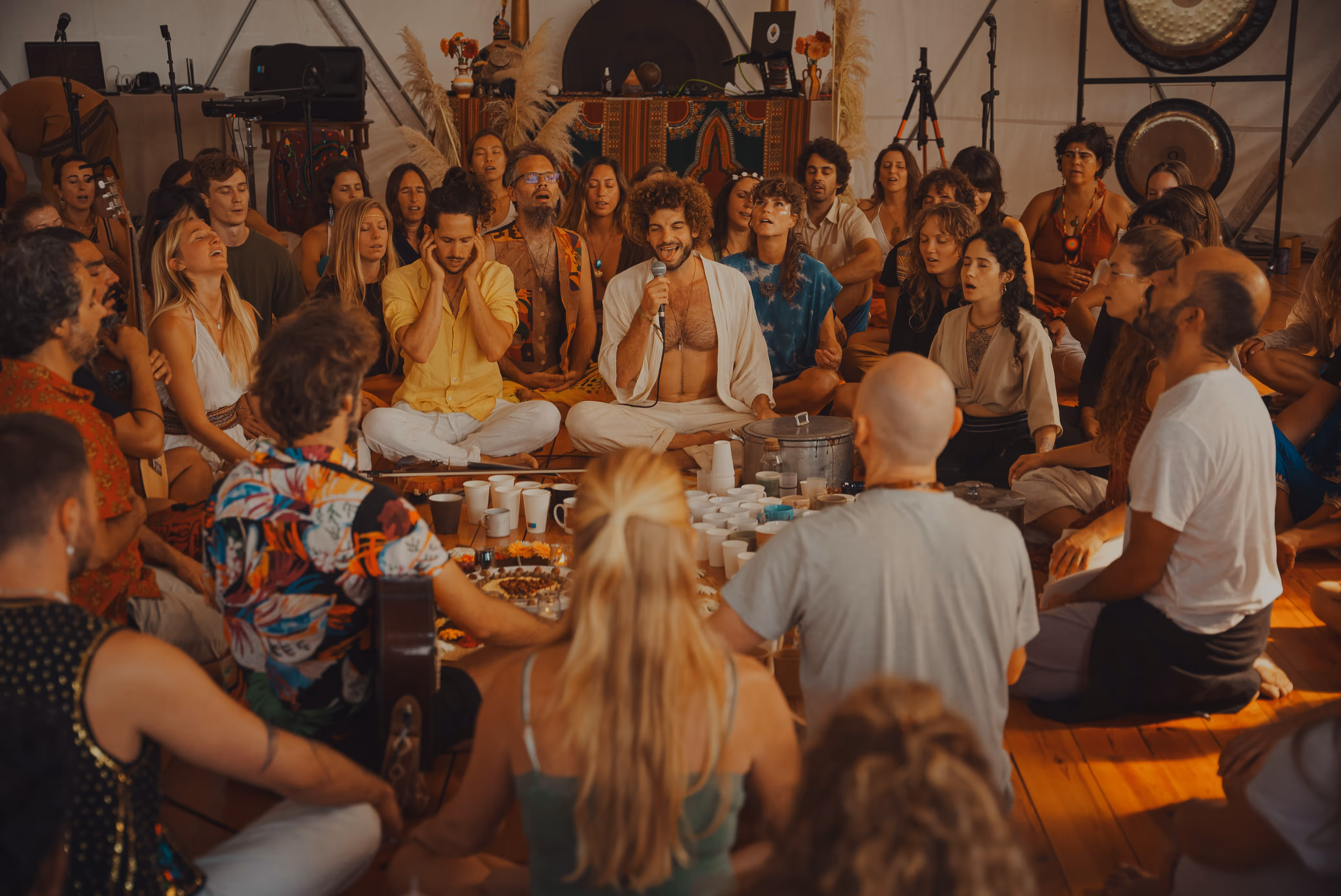 People sitting in a large circle on the floor, meditating and singing together in a warm, cozy room with musical instruments in the background.