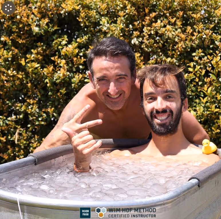 Two smiling men in an ice bath tub outdoors, one making a peace sign and a yellow rubber duck on the tub edge.