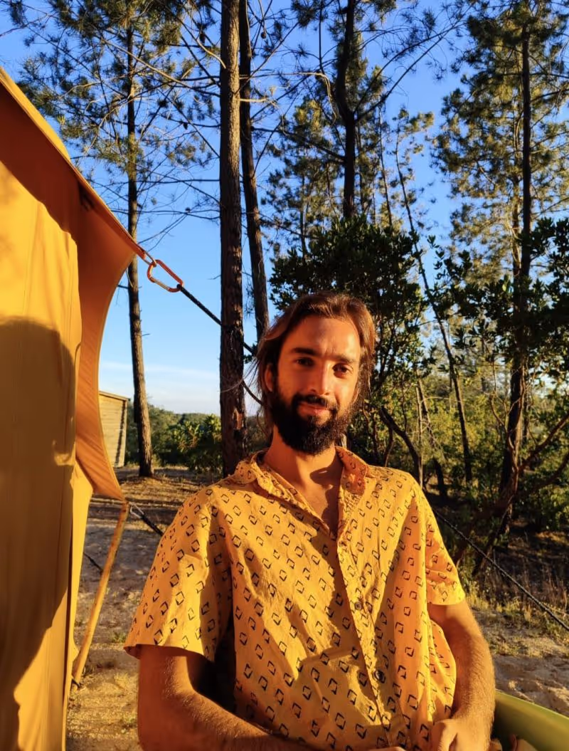 Man with beard sitting outdoors in sunlight wearing a yellow patterned shirt with trees and a tent in the background.