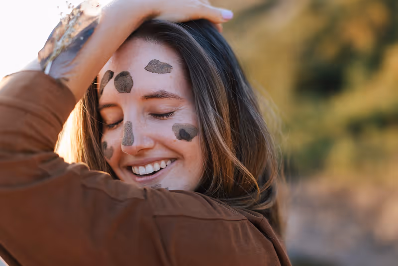 Smiling woman with closed eyes and dark facial mud patches, wearing a brown top, outdoors with blurred greenery background.